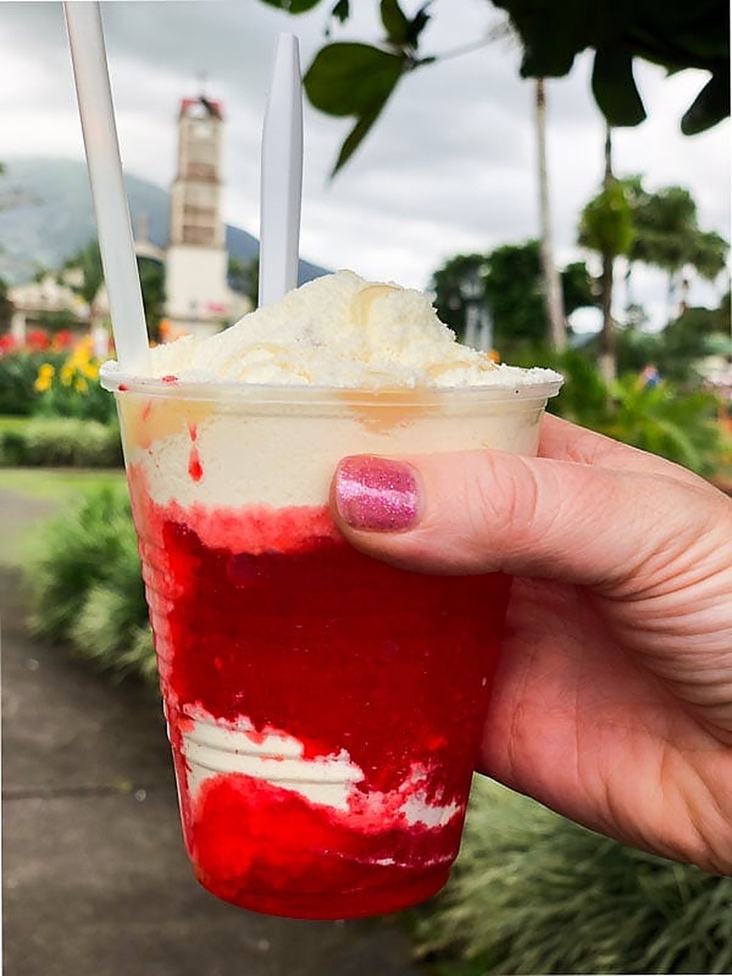 A real Costa Rican granizado with red fruit syrup and powdered milk, held in a park with a church tower behind