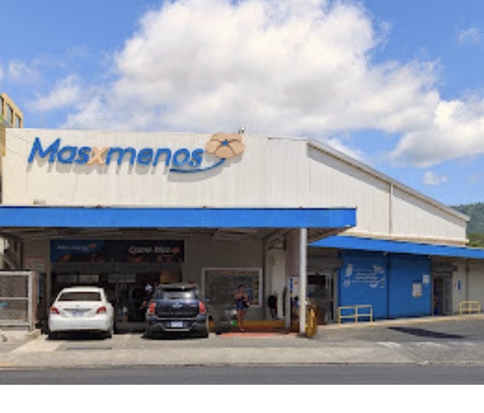 Storefront of the Más x Menos supermarket in Jacó on a sunny day, cars parked under the entrance canopy
