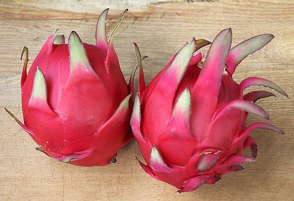 Two whole bright-pink dragon fruits with green tips resting on a wooden board